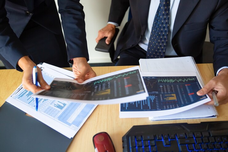 Two men discussing financial charts and data, holding papers and a pen.